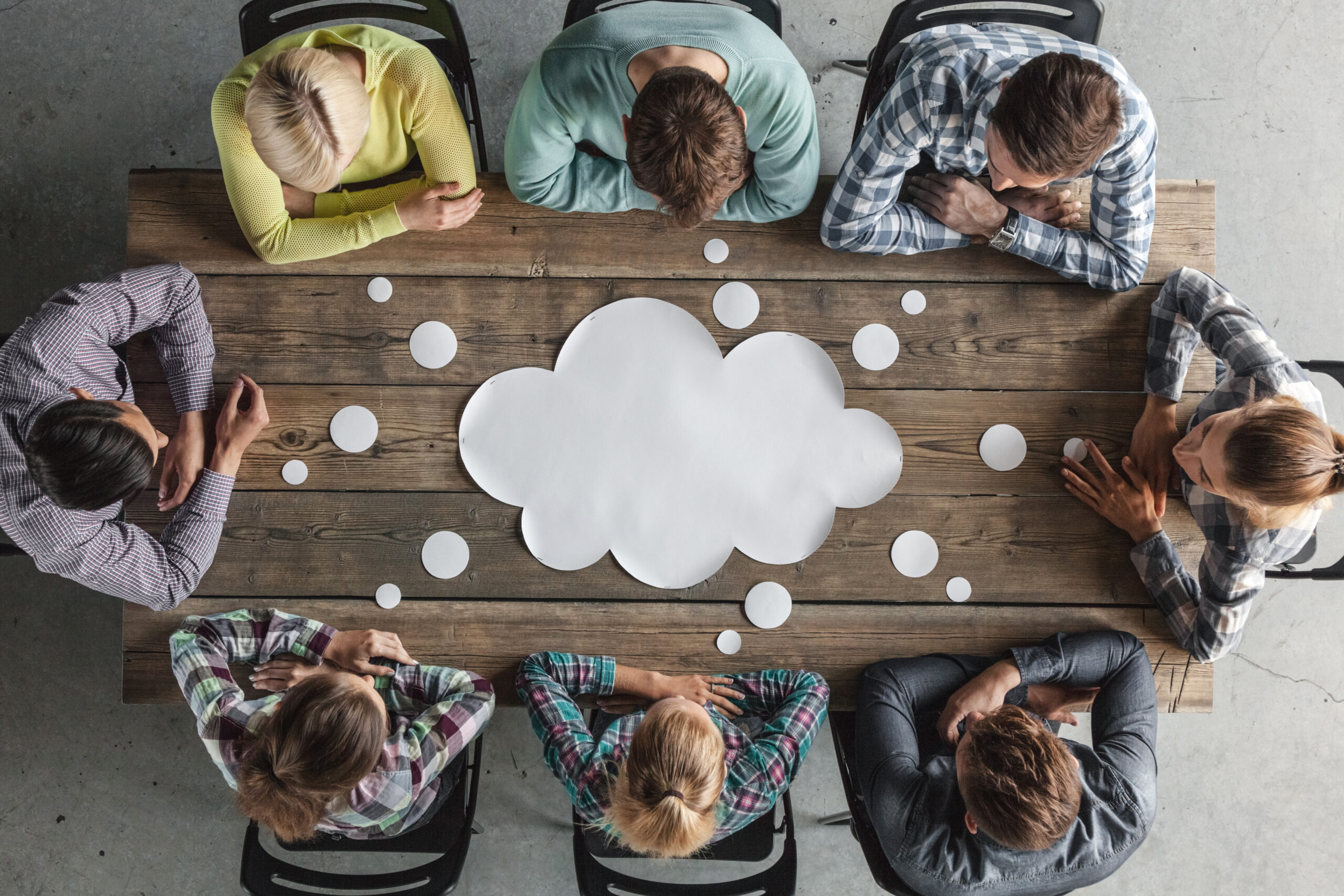 innovative technology solutions Eight people sit around a wooden table with a large paper cloud and small circles in the center, suggesting an agile development brainstorming session focused on innovative technology solutions. IT services